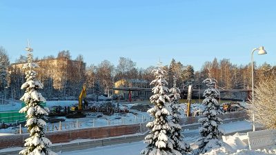 Talvinen maisema, taustalla rakennuksia, keskellä aidoin rajattu työmaa, jolla työmaakoneita.