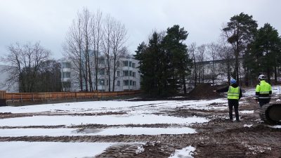 Kaksi miestä seisoo tasaisella kentällä työmaa-aitojen sisällä. Aidan ulkopuolella on kerrostaloja.