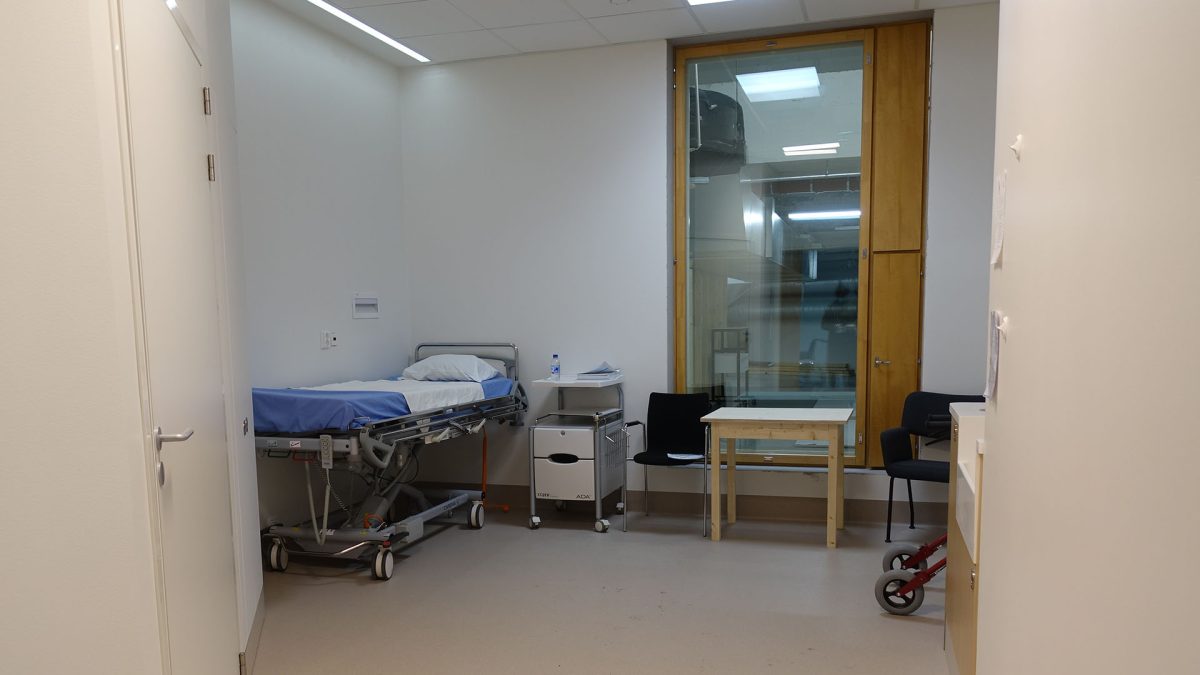 Sairaalahuoneessa on sänky nurkassa, yöpöytä, kaksi tuolia ja pieni irtopöytä sekä korkea ikkuna.