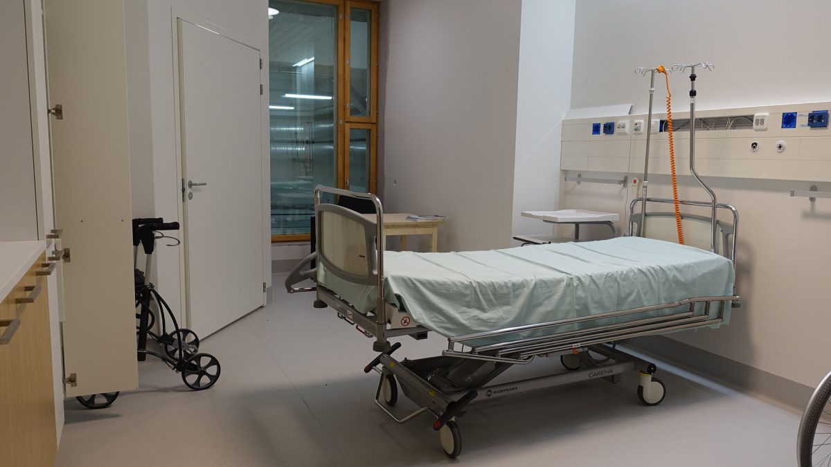 Sairaalasänky seisoo keskellä huonetta, toisella seinustalla on rollaattori.