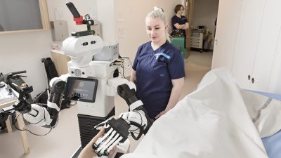 Sairaanhoitaja ja robotti seisovat potilassängyn vieressä potilashuoneessa.