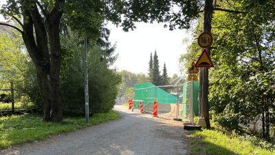 Vihreällä maalattu työmaa-aita ja punakeltaisia lamelleja on kauniin vihreän pyörätien reunassa.