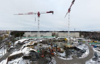 Kaksi korkeaa nosturia työmaa-alueella, taustalla kaksi isoa sairaalarakennusta.